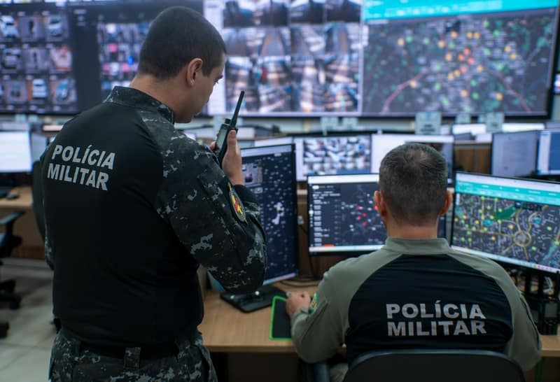 Policiais militares de Goiás em central de monitoramento com telões de câmeras e mapas do programa IA Contra o Crime